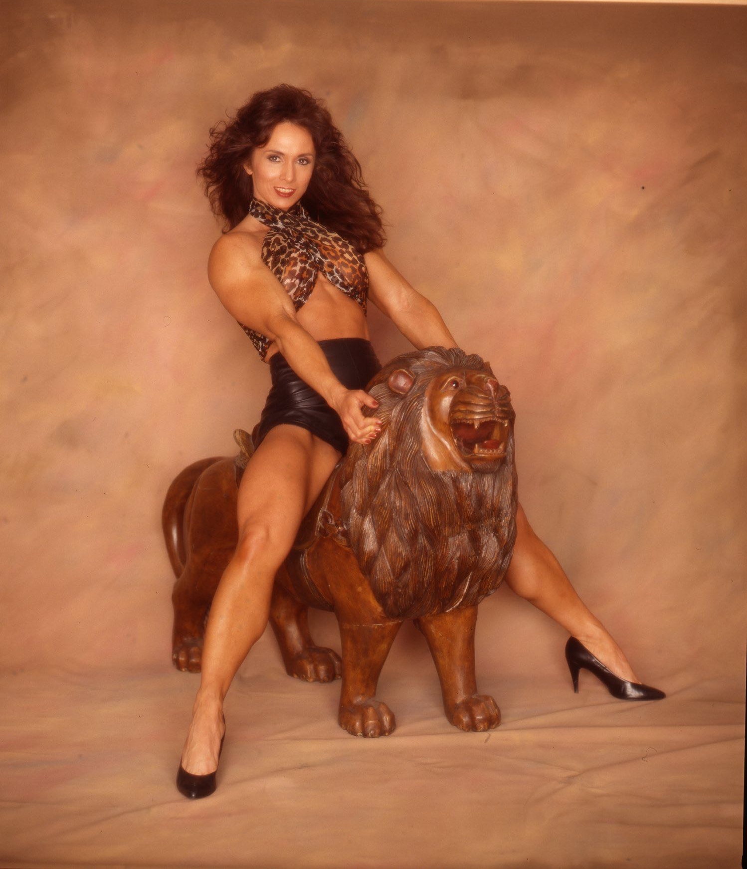 Deb McKnight IFBB Pro in black shorts and leopard top posing on top of a decorative lion at a photo shoot.