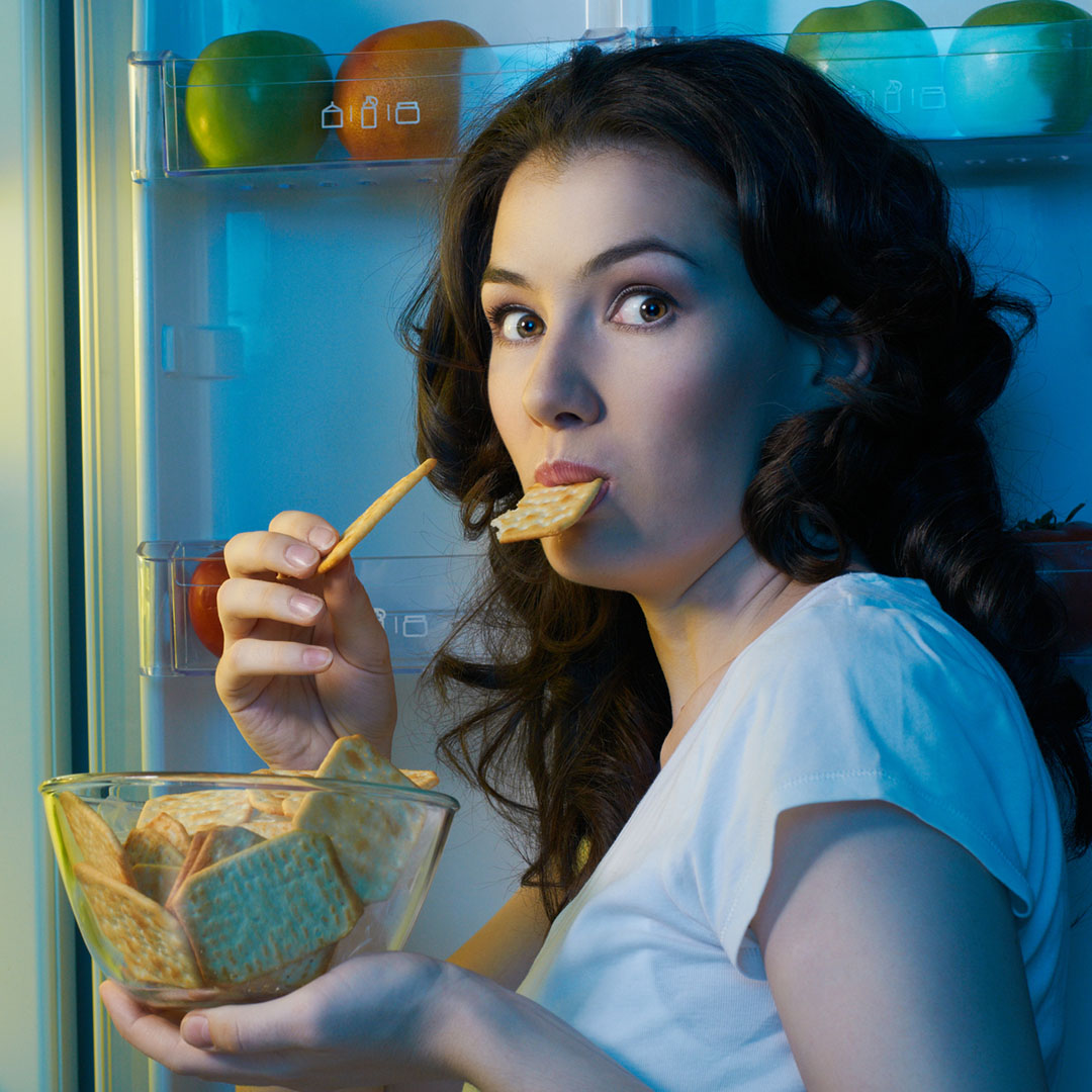 Young woman standing with open fridge late at night snacking on junk food