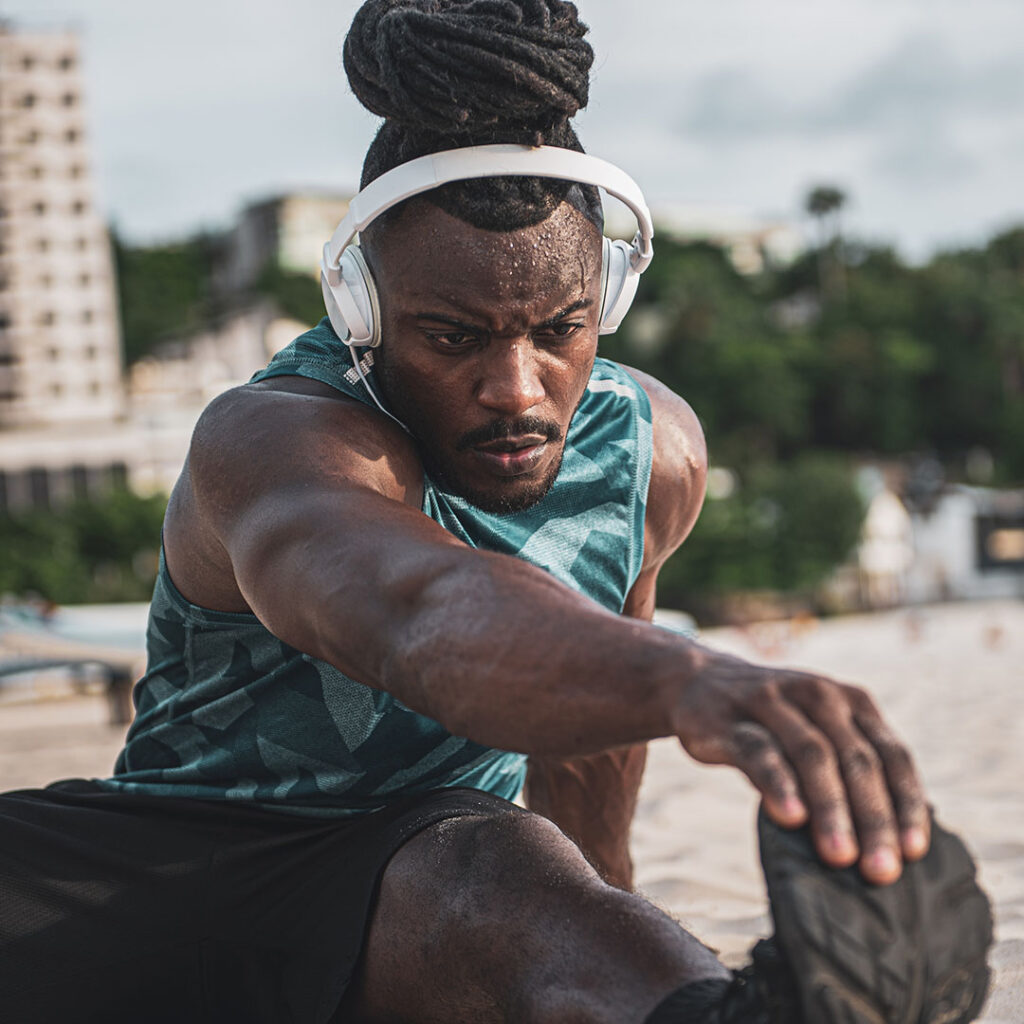 Athletic dark skinned man with headphones stretching his hamstrings on the beach