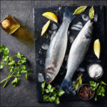 2 whole fresh Branzino on black cutting board on concrete table with sprigs of parsley and lemon